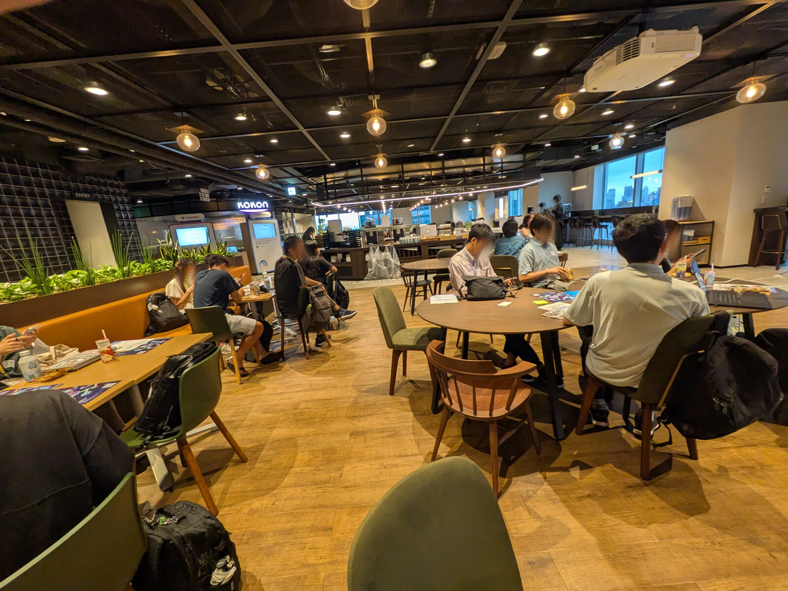 カフェスペースに置かれた丸テーブルを学生が囲み、開演前に資料を広げている様子。奥にはGeminiのサイネージと窓の外の夕景が見える